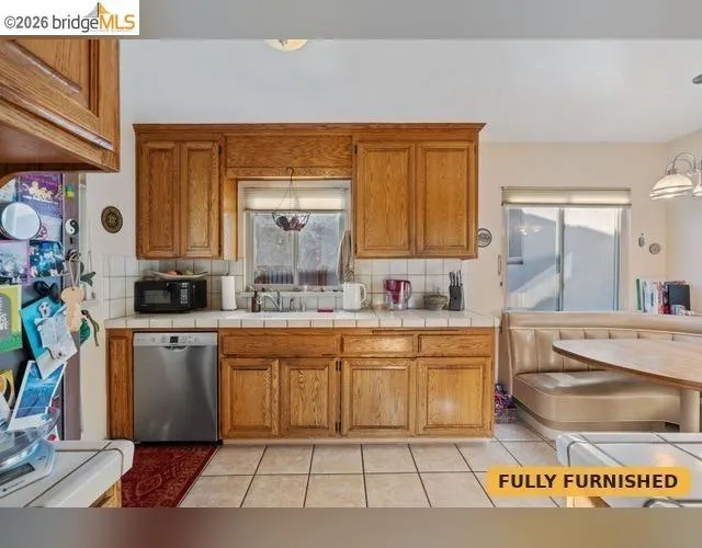 Wood-finish cabinetry, tile countertops, stainless steel dishwasher, and tile flooring