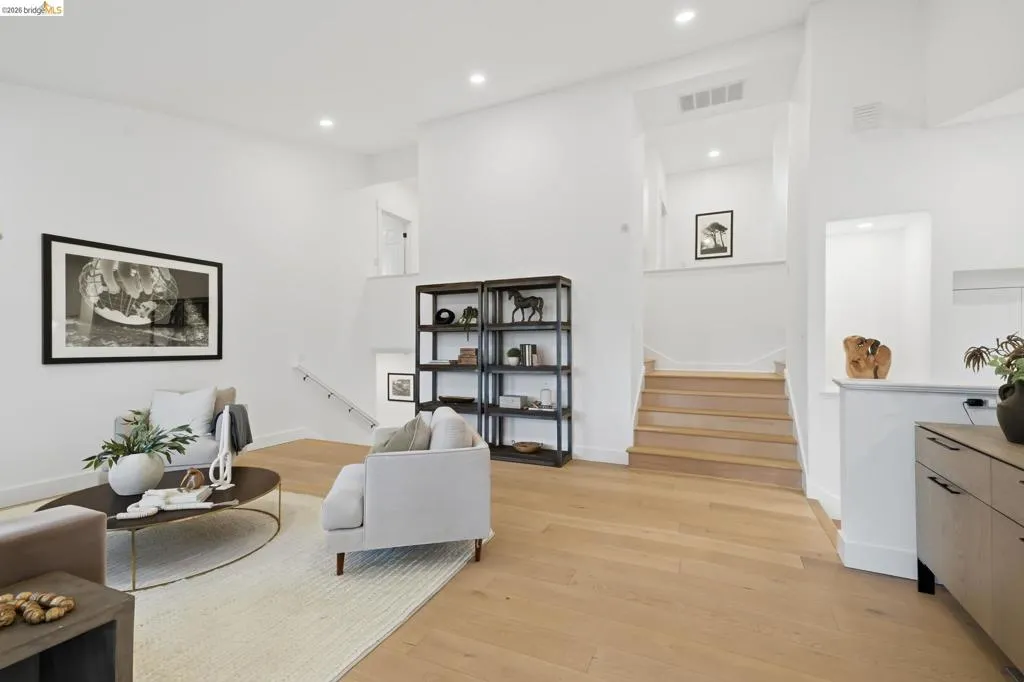 Open-concept living area featuring light wood-finish flooring, recessed lighting, a built-in shelving unit, and a staircase with a minimalist railing Open-concept living area featuring light wood-finish flooring, recessed lighting, a built-in shelving unit, and a staircase with a minimalist railing