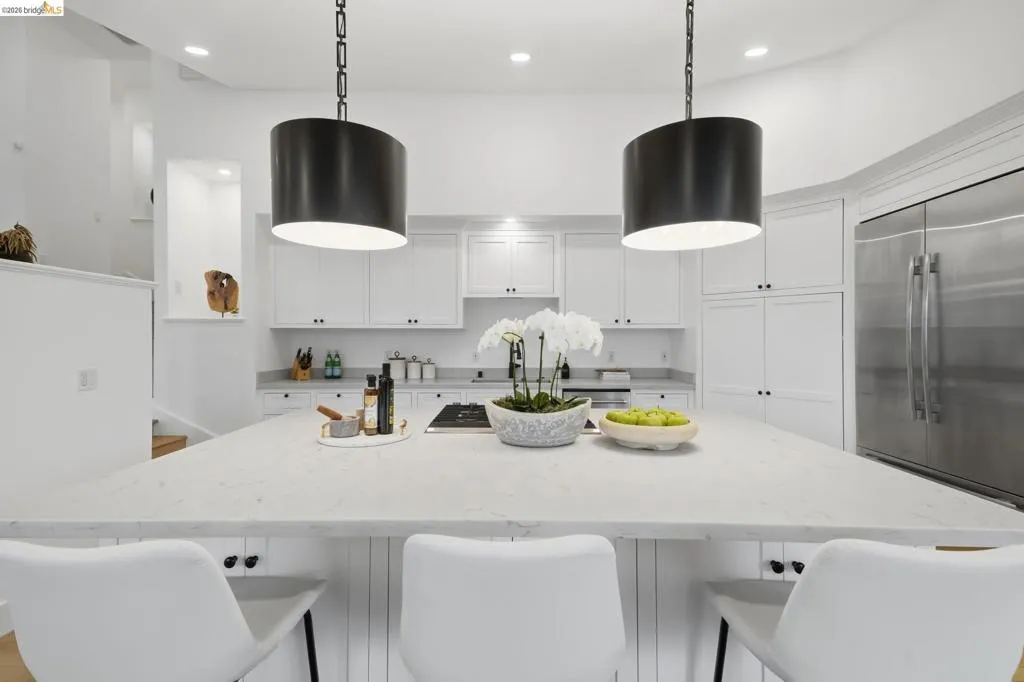 Expansive kitchen island with a light-toned countertop, white cabinetry with dark hardware, stainless steel refrigerator, and recessed lighting Expansive kitchen island with a light-toned countertop, white cabinetry with dark hardware, stainless steel refrigerator, and recessed lighting