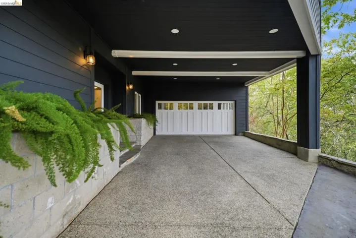 Covered carport with exposed aggregate concrete drive, white paneled garage door, dark exterior siding, and integrated wall sconces