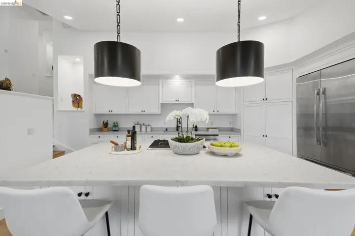 Expansive kitchen island with a light-toned countertop, white cabinetry with dark hardware, stainless steel refrigerator, and recessed lighting
