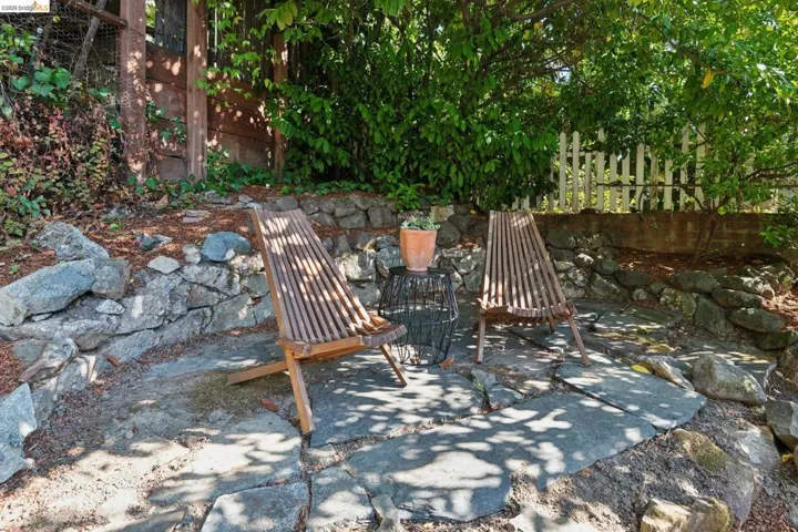 Stone patio area featuring natural rock retaining walls, mature green foliage, and a white picket fence