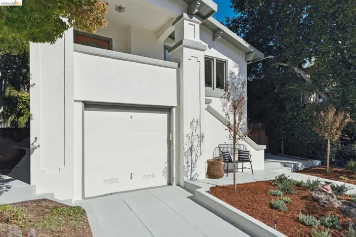 White stucco exterior with a garage, concrete driveway, front porch, mature trees, and landscaped garden beds