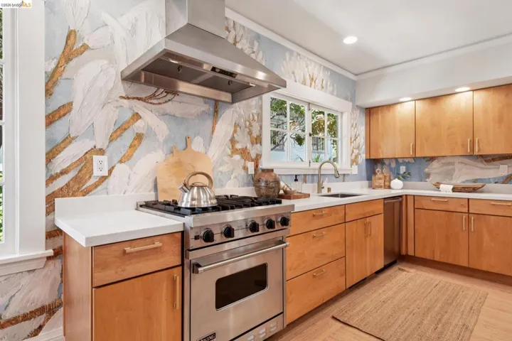 Built-in stainless steel range with overhead ventilation, light wood-finish cabinetry, white countertops, and a stainless steel sink with a gooseneck faucet