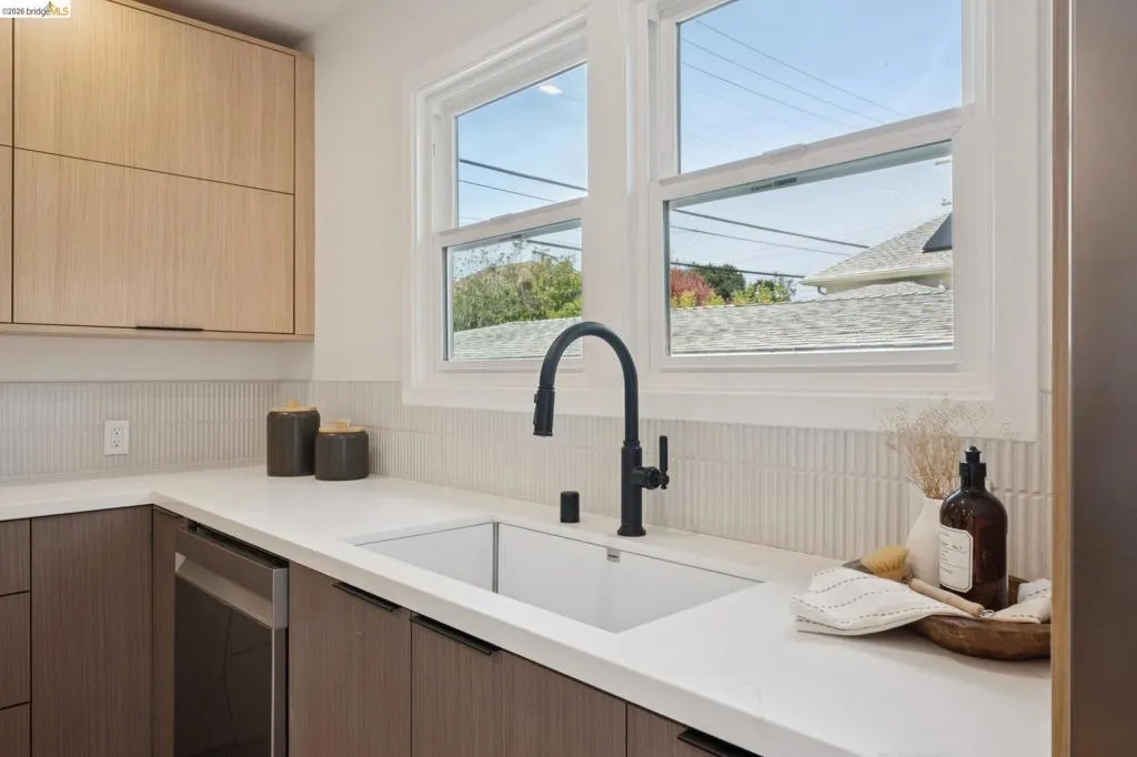 Integrated stainless steel dishwasher, white countertop, undermount sink with a matte black faucet, wood-finish lower cabinetry, and light wood-finish upper cabinetry