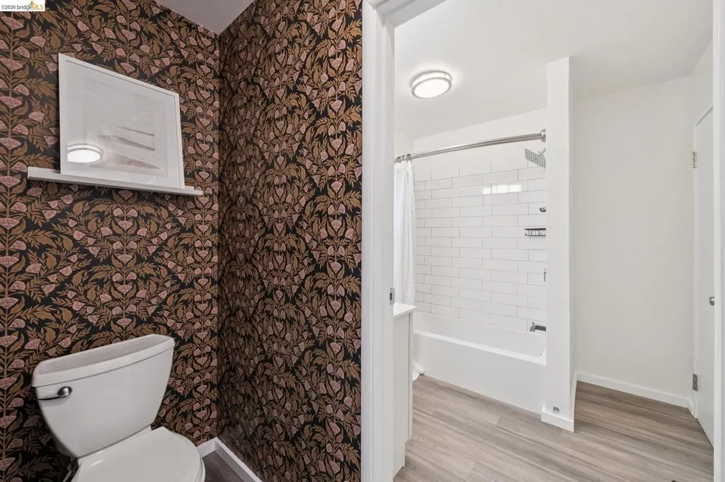 Patterned wallpaper, white wall shelving, water closet, wood-finish flooring, and a subway tile shower surround
