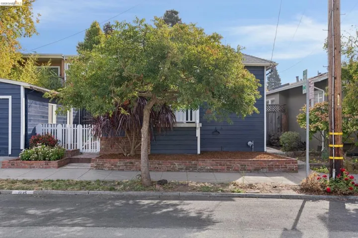 Dark blue horizontal siding exterior with a white picket fence, brick planter boxes, mature tree, and established landscaping