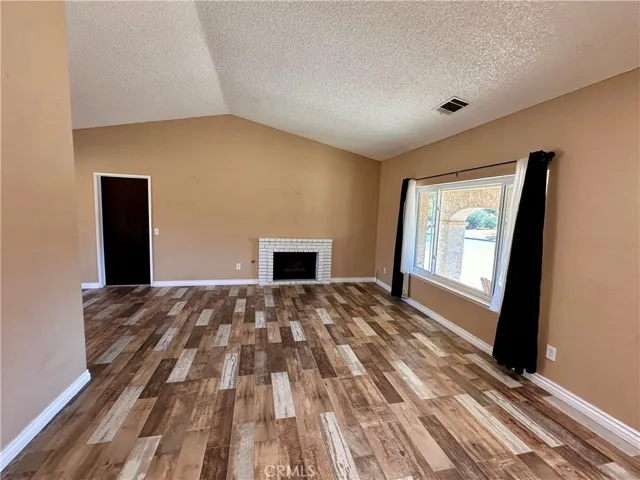 This spacious living room features vaulted ceilings, wood-look flooring, a white brick fireplace, and a large front-facing window that brings in abundant natural light.