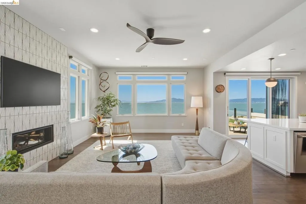 Expansive water scenery visible through multiple windows and sliding glass doors, built-in fireplace with a decorative tile surround, recessed lighting, and dark wood-finish flooring