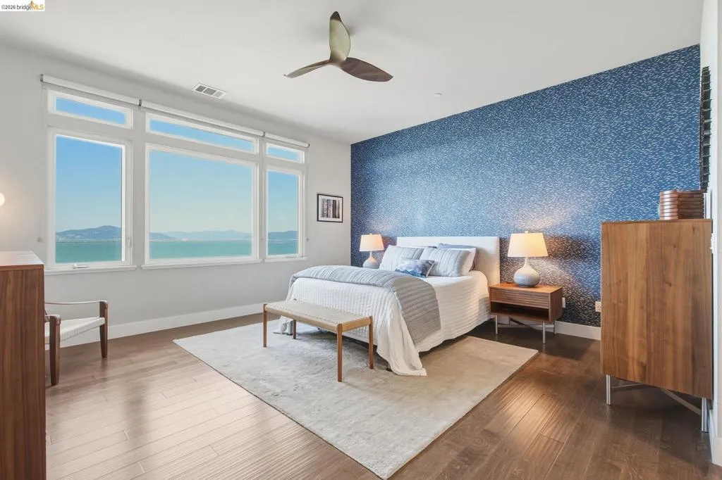 Expansive window wall with water scenery, wood-finish flooring, accent wall with textured wallpaper, and a ceiling fan