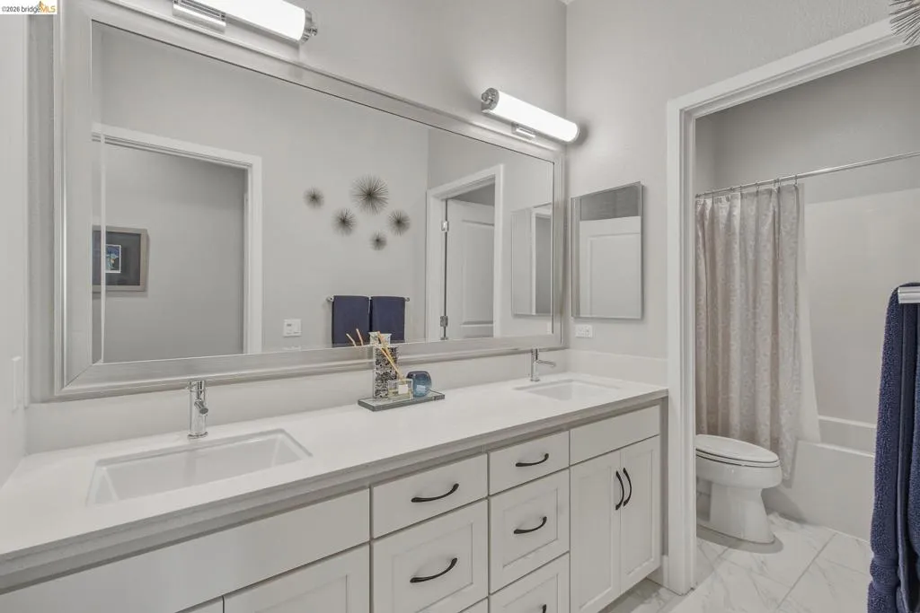 Dual undermount sinks with chrome fixtures, white shaker-style cabinetry, a large framed vanity mirror, and an adjacent shower-tub combination