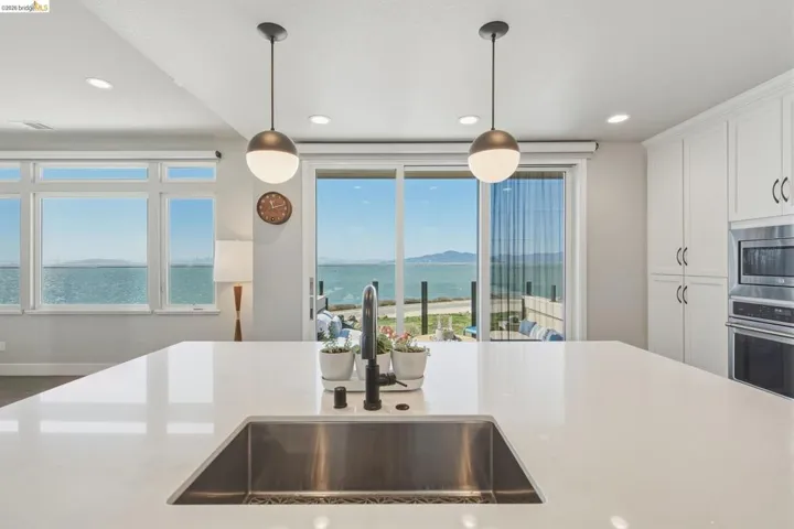 Expansive kitchen island with an undermount stainless steel sink, white cabinetry, integrated stainless steel appliances, and a sliding glass door to an outdoor area