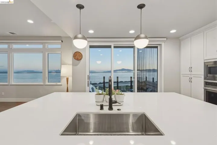 Expansive island with integrated stainless steel sink, sleek pendant lighting, white cabinetry, built-in stainless steel appliances, and large windows with water scenery
