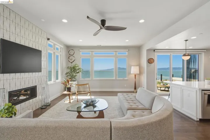 Expansive water scenery visible through multiple windows and sliding glass doors, built-in fireplace with a decorative tile surround, recessed lighting, and dark wood-finish flooring