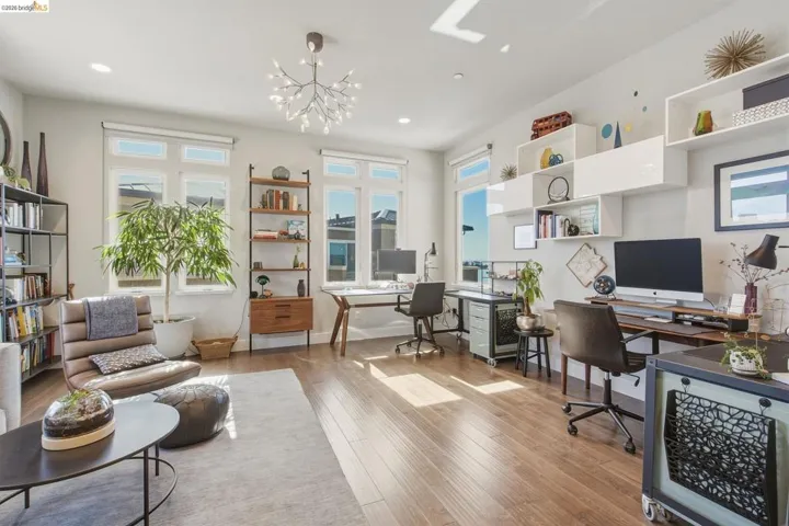 Bright, open living space featuring hardwood flooring, abundant natural light from multiple windows, built-in shelving with display storage, a modern pendant light fixture, and a dedicated work station with integrated desk cabinetry