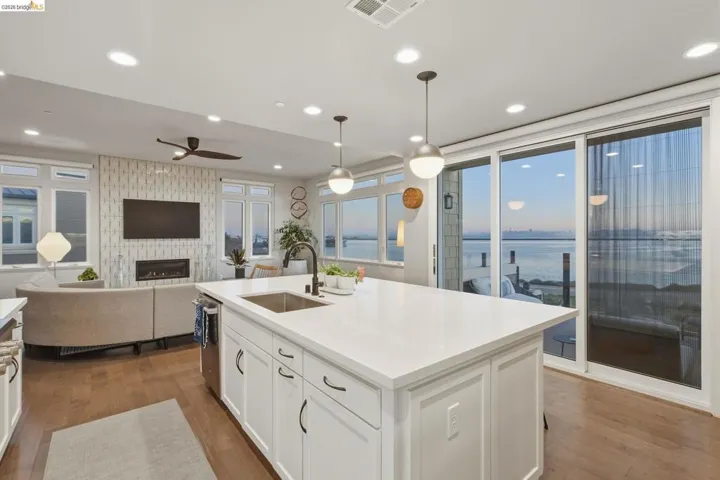 Open-concept living space with wood-finish flooring, a contemporary fireplace feature wall, a spacious kitchen island with an undermount sink, pendant lighting, and expansive sliding glass doors