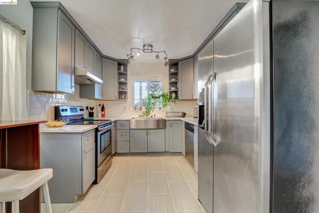Kitchen featuring gray cabinetry, stainless steel appliances, white subway tile backsplash, light-toned tile flooring, and a farmhouse sink