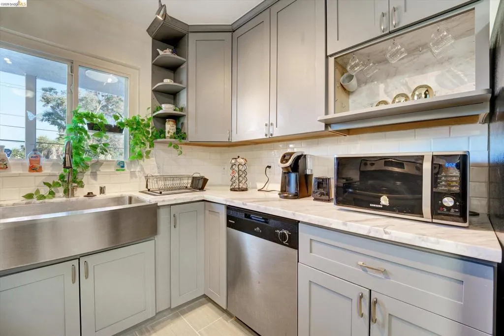 Stainless steel apron-front sink, light gray cabinetry, white subway tile backsplash, light-toned countertop, and stainless steel dishwasher