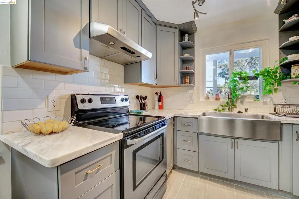 Gray cabinetry, white subway tile backsplash, white countertops, stainless steel farmhouse sink, and stainless steel range with overhead ventilation