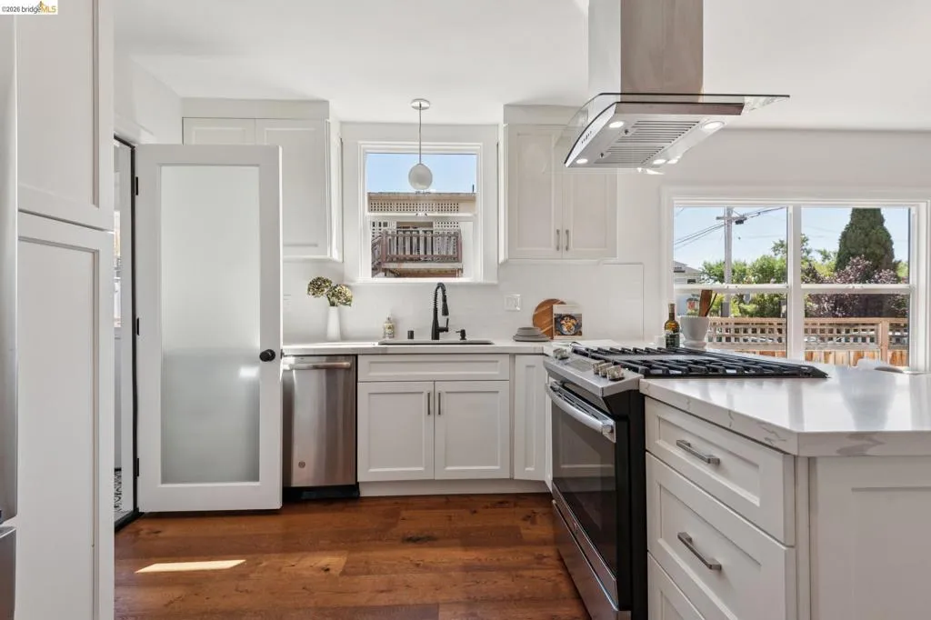 White shaker cabinetry with ample storage, stainless steel appliances, a modern range hood, expansive countertops, and wood-finish flooring White shaker cabinetry with ample storage, stainless steel appliances, a modern range hood, expansive countertops, and wood-finish flooring