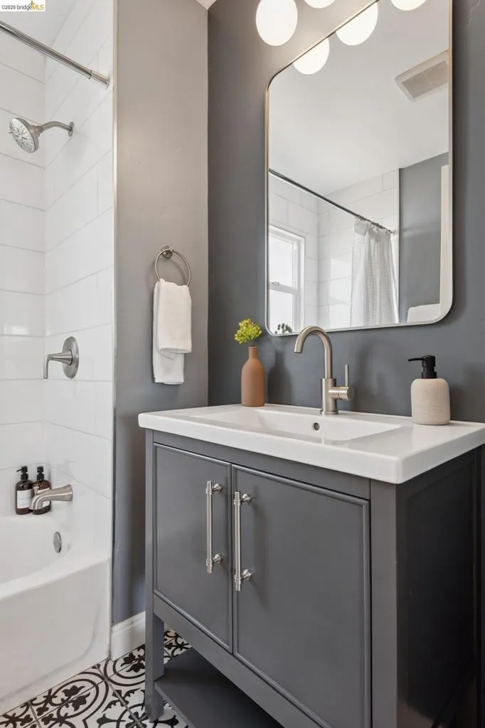Bathroom featuring a grey vanity with a white countertop, patterned floor tiles, white subway tile shower surround, and a wall-mounted mirror with integrated lighting Bathroom featuring a grey vanity with a white countertop, patterned floor tiles, white subway tile shower surround, and a wall-mounted mirror with integrated lighting