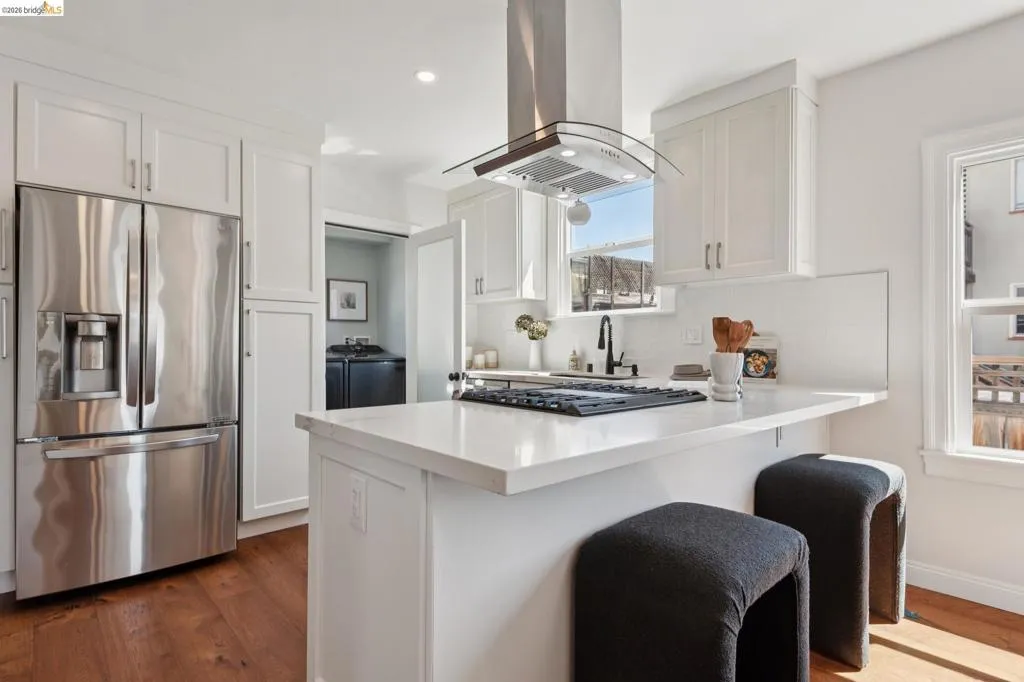 Stainless steel French door refrigerator, white shaker-style cabinetry, wood-finish flooring, white countertop island, and stainless steel range hood Stainless steel French door refrigerator, white shaker-style cabinetry, wood-finish flooring, white countertop island, and stainless steel range hood