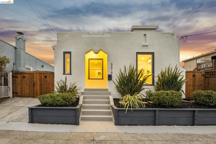 Stucco facade with decorative entrance archway, dark-trimmed windows, concrete steps, and front yard landscaping with raised planters
