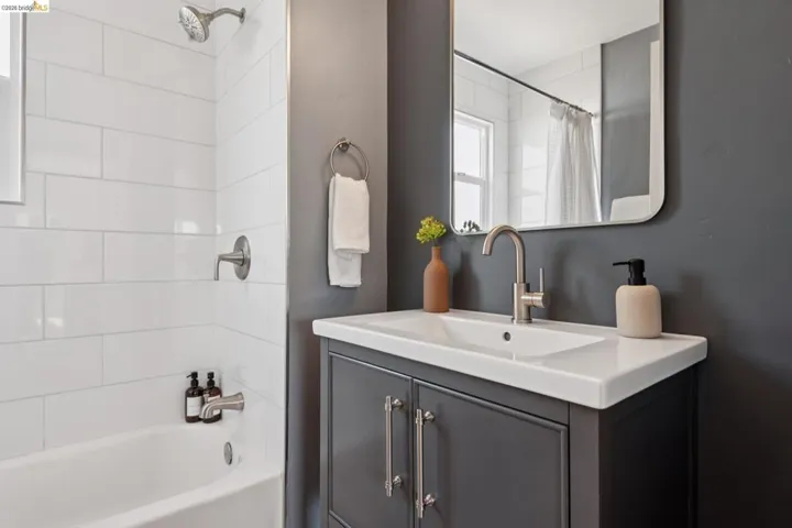 White subway tile shower surround, bathtub with brushed nickel fixtures, dark gray vanity with integrated white sink, brushed nickel faucet, and a rectangular framed mirror