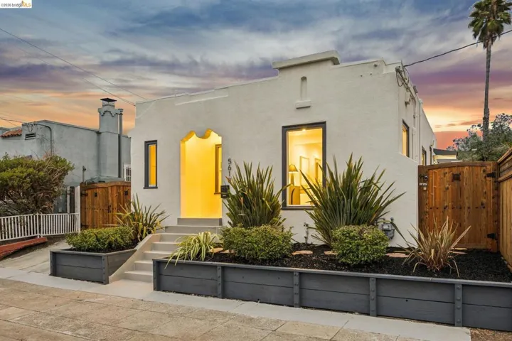 Stucco exterior with arched entryway, dark-framed windows, concrete steps, and raised garden beds with planted shrubs