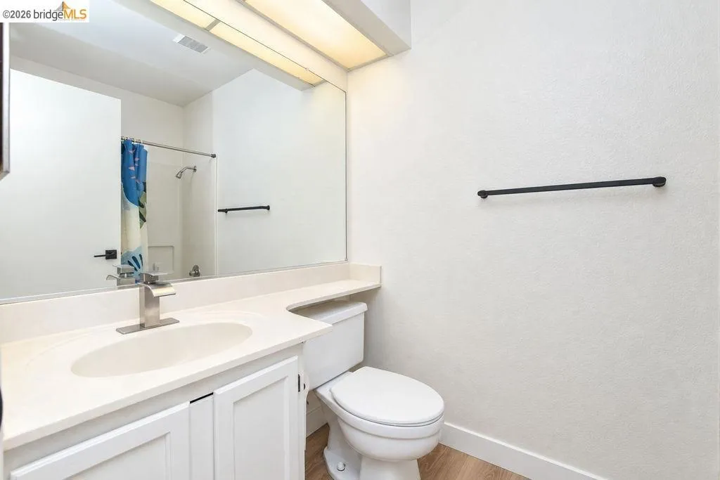 Vanity with an integrated sink and brushed nickel faucet, large wall-mounted mirror, white shaker-style cabinetry, and wood-finish flooring
