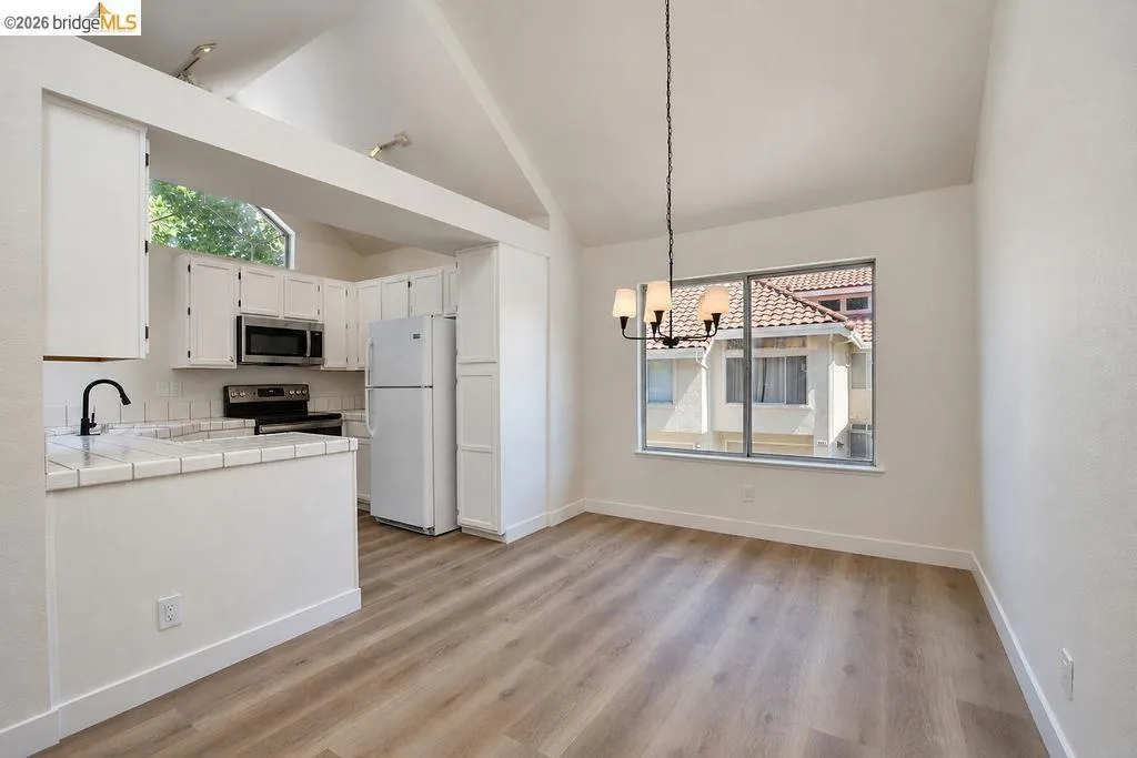 Vaulted ceiling, wood-finish flooring, white cabinetry, tile countertops, and a window with a neighborhood backdrop