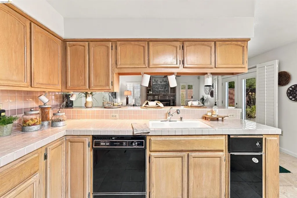 Wood cabinetry, tile countertops, tile backsplash, built-in dishwasher, and a pass-through opening