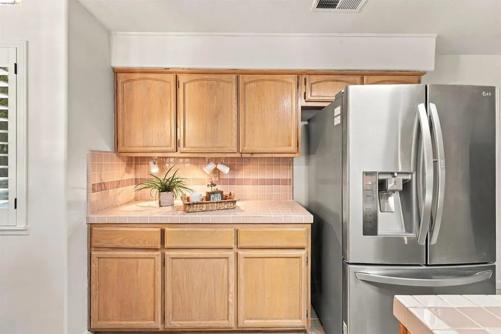 Wood-finish cabinetry, tile countertops, tile backsplash, and a stainless steel refrigerator
