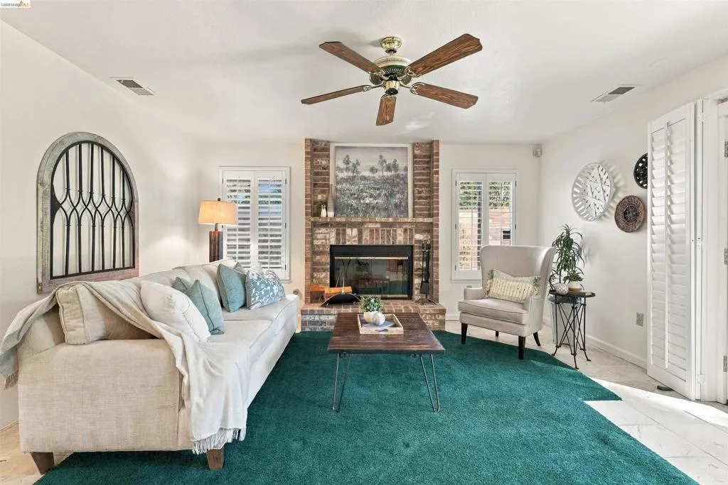 Brick fireplace with a black firebox, white plantation shutters, and a brass-finish ceiling fan