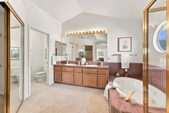 Vaulted ceiling, dual vanity, built-in jetted tub, wood-finish cabinetry, and mirrored closet doors