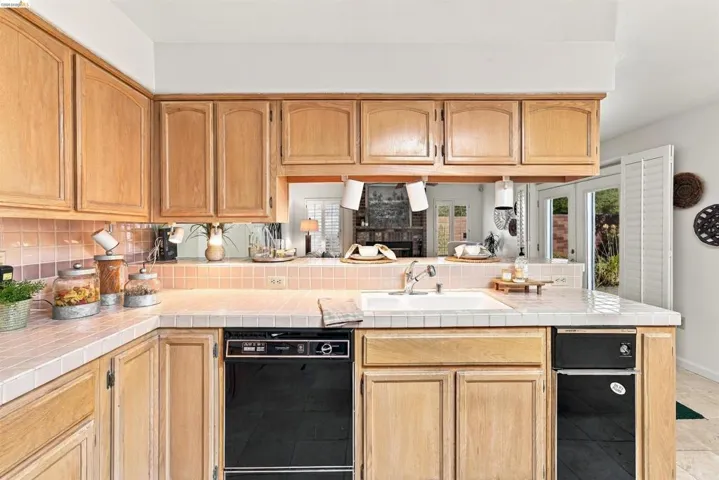 Wood cabinetry, tile countertops, tile backsplash, built-in dishwasher, and a pass-through opening