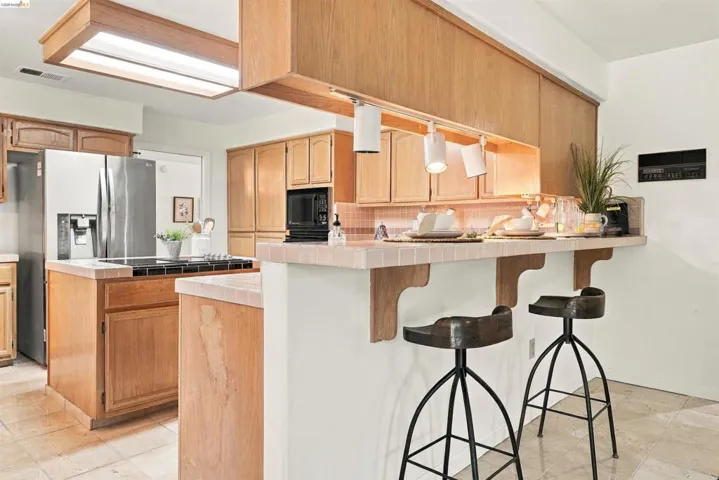 Wood-finish cabinetry, tile countertops, built-in microwave, stainless steel refrigerator, and tiled flooring