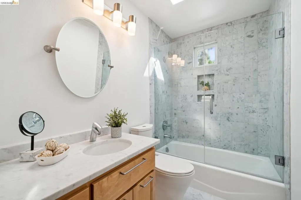 Bathroom featuring a vanity with a white stone countertop, a glass-enclosed tub and shower with tile surround, and a skylight Bathroom featuring a vanity with a white stone countertop, a glass-enclosed tub and shower with tile surround, and a skylight