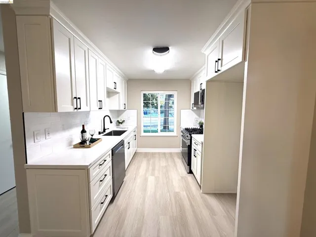 Galley kitchen featuring white shaker cabinetry, light wood-finish flooring, a white tiled backsplash, stainless steel appliances, and a window