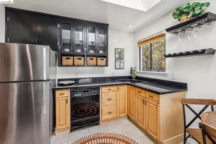 Stainless steel refrigerator, black upper cabinetry with glass inserts, light wood-finish lower cabinetry, dark stone-finish countertops, and a built-in dishwasher