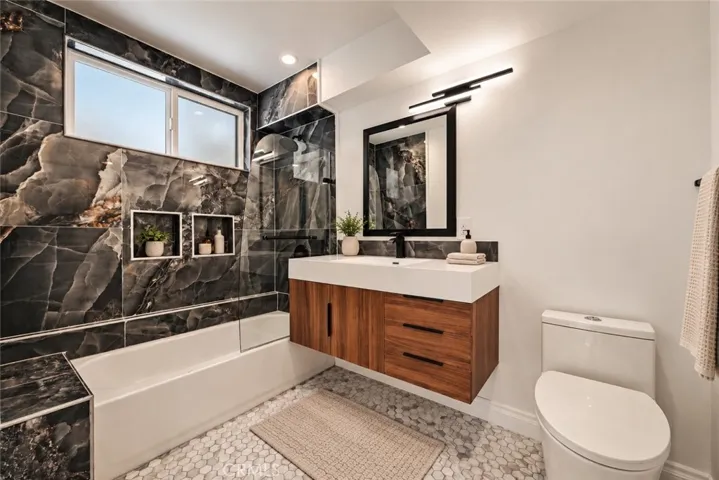 The primary suite features a walk-in closet and remodeled private bath with mosaic tile flooring, floating vanity, designer lighting, and custom-tiled tub/shower combination with glass panel. Photo is virtually staged.