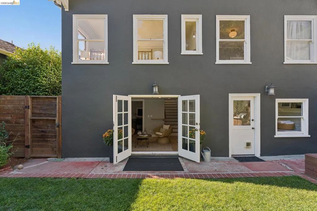 Exterior rear facade featuring smooth dark-toned stucco, white-framed multi-pane French doors, a manicured lawn, and a brick-bordered concrete patio