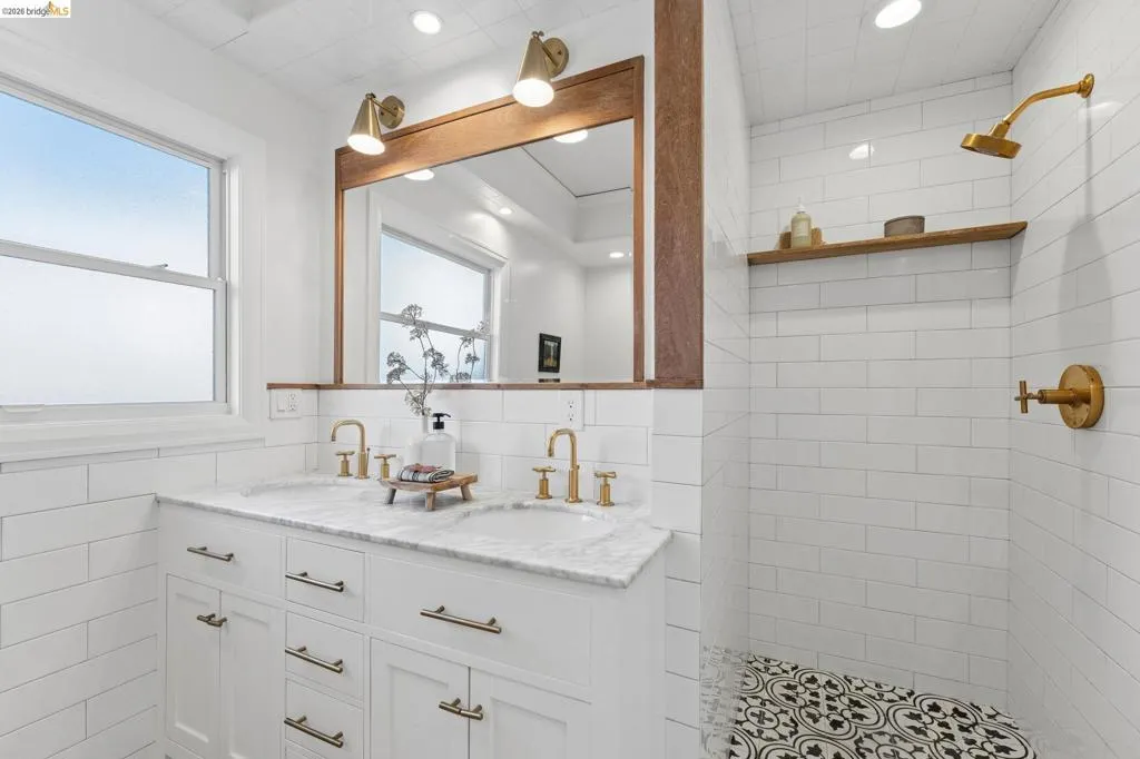Dual-sink vanity with stone countertop, wood-framed mirror, brass-finish fixtures, subway tile wall cladding, and patterned floor tile
