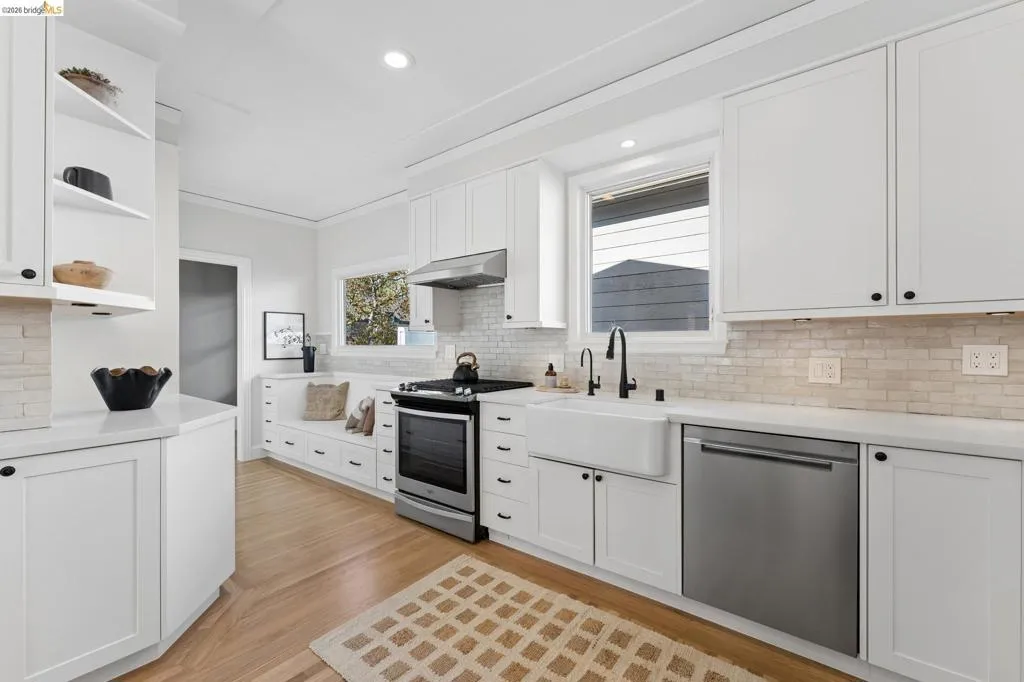 Kitchen featuring white shaker-style cabinetry, a porcelain farmhouse sink with matte black hardware, light-toned stone backsplash, stainless steel appliances, and integrated bench seating with built-in storage