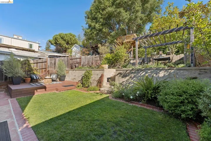 Multilevel outdoor space featuring a wood-finish deck, tiered brick-edged landscaping, a concrete retaining wall with integrated stone steps, and a wooden pergola