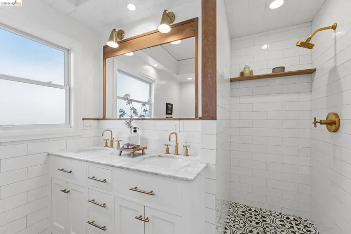 Dual-sink vanity with stone countertop, wood-framed mirror, brass-finish fixtures, subway tile wall cladding, and patterned floor tile