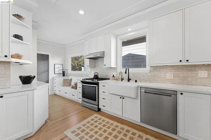 Kitchen featuring white shaker-style cabinetry, a porcelain farmhouse sink with matte black hardware, light-toned stone backsplash, stainless steel appliances, and integrated bench seating with built-in storage