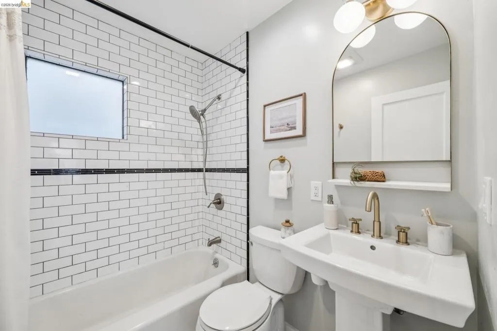 Bathroom featuring white subway tile shower surround with black accent border, pedestal sink with brass-finish faucet, arched mirror, and brass-finish multi-light wall fixture