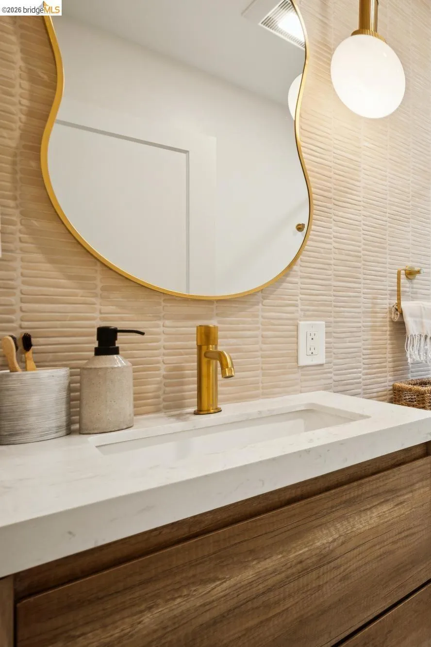 Floating wood-finish vanity with a rectangular integrated sink, stone countertop, brass-finished faucet, textured wall tile, and an organic-shaped brass-trimmed mirror