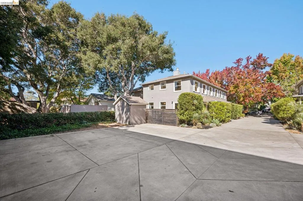Extensive concrete motor court featuring mature trees, landscaped shrubbery, a detached storage structure, and horizontal wood-slat fencing